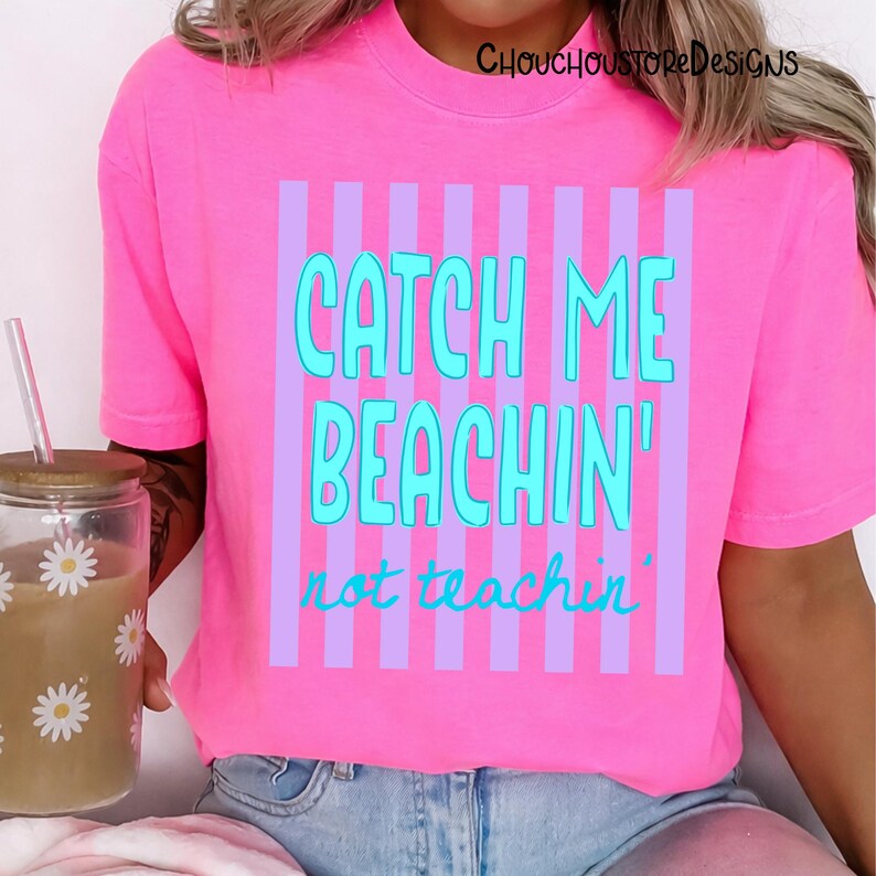 May include: A bright pink t-shirt with the text "CATCH ME BEACHIN' not teachin'" in turquoise and purple. The shirt has vertical purple stripes behind the text. The shirt is a casual wear item.