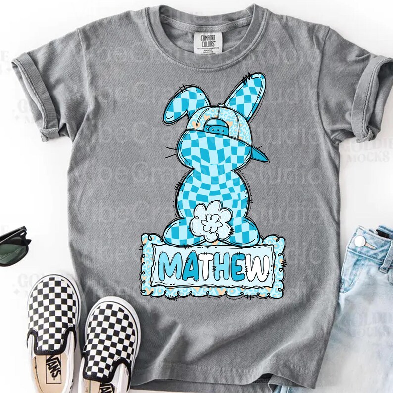 May include: Gray t-shirt featuring a cartoon bunny wearing a baseball cap with a blue checkered pattern. The bunny is holding a flower and the name "MATHEW" is displayed on a blue sign. Black and white checkered Vans shoes are visible.