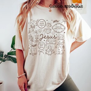 May include: Cream-colored t-shirt featuring a black and white graphic design with the word "Jesus" in the center. The design includes religious symbols, text, and illustrations such as a cross, a lamb, and the phrases "Light of the world" and "Grace Upon Grace."