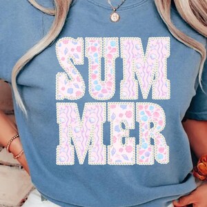 May include: A light blue t-shirt with the word "SUMMER" in large, decorative letters. The letters are filled with a pattern of seashells and other summer-themed designs in pastel colors. The shirt is a casual style.