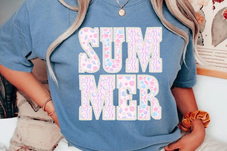 May include: A light blue t-shirt with the word "SUMMER" in large, decorative letters. The letters are filled with a pattern of seashells and other summer-themed designs in pastel colors. The shirt is a casual style.