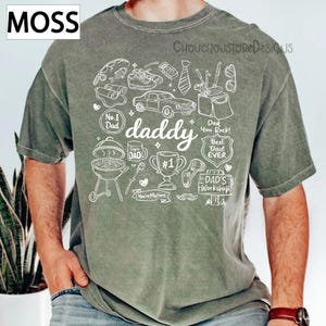 May include: A moss green t-shirt with a white graphic design featuring the word "daddy" and various illustrations like a car, trophy, and barbecue. The shirt also includes the text "Best Dad Ever" and "Dad You Rock!"