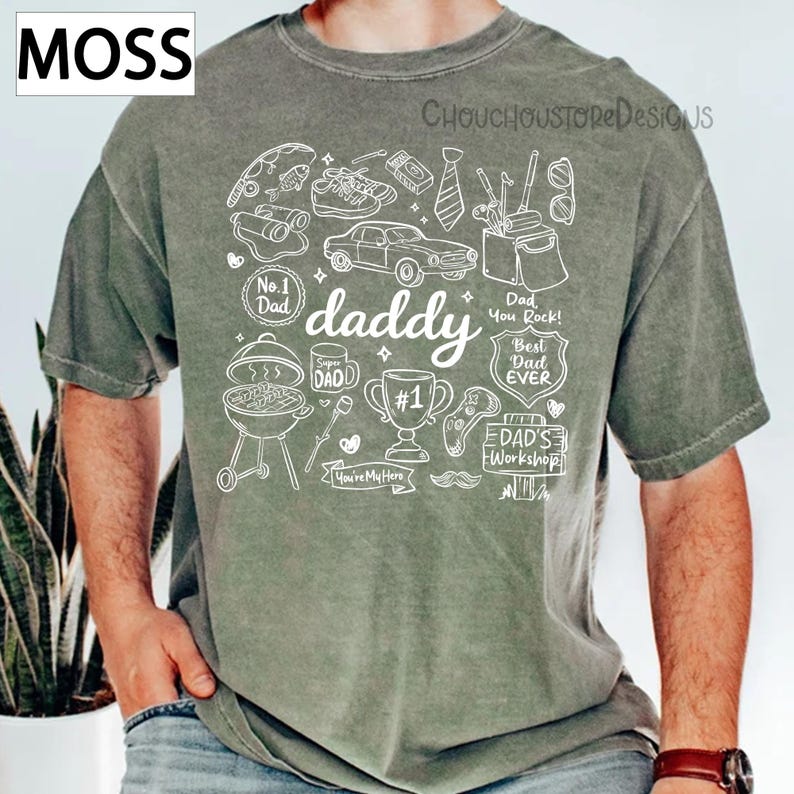 May include: A moss green t-shirt with a white graphic design featuring the word "daddy" and various illustrations like a car, trophy, and barbecue. The shirt also includes the text "Best Dad Ever" and "Dad You Rock!"