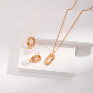 May include: A gold necklace and earrings set with a delicate chain and a pendant featuring a geometric design with a pearl accent.