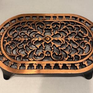 May include: An oval-shaped, cast iron trivet with an ornate, openwork design. The trivet features a copper-colored finish on the outer rim and decorative elements, with a black interior. It has small feet and a handle on one side.