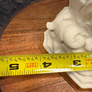 May include: A cream-colored ceramic figurine with gold trim, measuring approximately 4 inches (10 cm) in width. The figurine has a detailed, flowing design. A yellow measuring tape is placed across the base of the figurine.