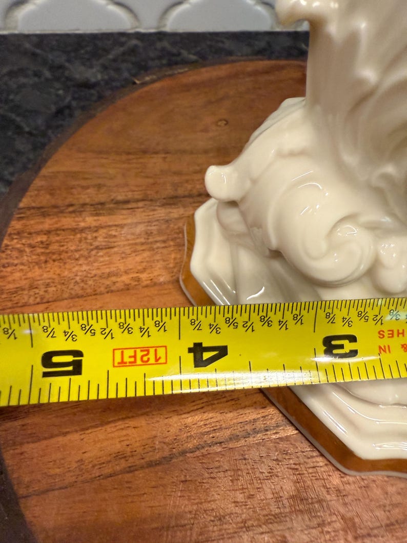 May include: A cream-colored ceramic figurine with gold trim, measuring approximately 4 inches (10 cm) in width. The figurine has a detailed, flowing design. A yellow measuring tape is placed across the base of the figurine.