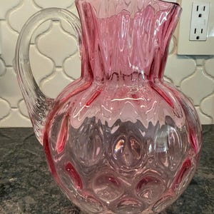 May include: A pink glass pitcher with a clear handle. The pitcher has a textured, bubble-like pattern on the bottom and a ruffled top. The handle is clear and curved. The pitcher is likely used for serving beverages.