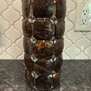 May include: A cylindrical vase constructed from dark brown, square coconut shell tiles. The tiles are arranged in a grid pattern and connected by thin, silver-colored metal wire loops at each corner, creating a textured, open-frame structure.