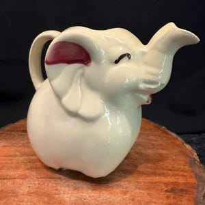 May include: A cream-colored ceramic pitcher shaped like an elephant. The pitcher features a curved handle on the back, a trunk forming the spout, and painted maroon accents on the inner ears. The elephant has closed eyes and a smooth, glossy finish.