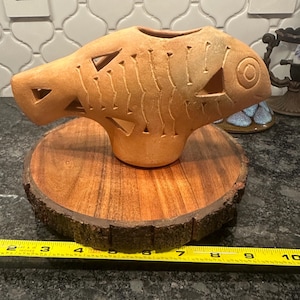 May include: A terracotta fish-shaped vase with intricate carvings and a hole in the top. The vase is sitting on a wooden slice.