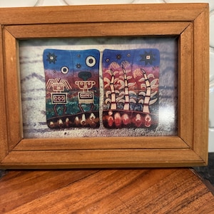 Puede incluir: Una obra de arte enmarcada con dos paneles abstractos. Los paneles muestran diseños coloridos sobre fondos azules y rojos, con formas geométricas. El marco es de madera.
