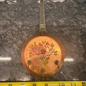 May include: A copper-colored wall-mounted key holder with a long handle and a round base. The base features a hand-painted floral design in red, pink, and blue. Three hooks are positioned below the floral design. The handle has decorative etching and a tag that reads "MADE IN SWITZERLAND".
