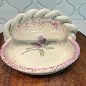May include: A cream-colored ceramic basket with a twisted handle. The bowl features a hand-painted purple and green floral motif at the center, with a speckled pink rim and accents along the edges of the bowl and the base of the handle.