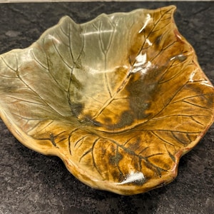 May include: A ceramic leaf-shaped bowl with a naturalistic design. The bowl features a blend of earthy tones, including olive green, brown, and golden hues. The intricate veining details add to its organic aesthetic. This decorative bowl is suitable for serving or display.