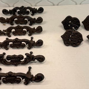 May include: A collection of six dark brown cast metal drawer pulls with ornate, scrolling designs arranged in a vertical column on the left, and four matching dark brown cast metal drawer knobs with leaf-like patterns arranged in a square on the right.