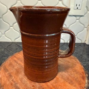 May include: A brown ceramic pitcher with a glossy finish, featuring a wide flared rim, a sturdy side handle, and a cylindrical body decorated with horizontal ribbed textures. The pitcher has a distinct pouring spout and a smooth, glazed surface.