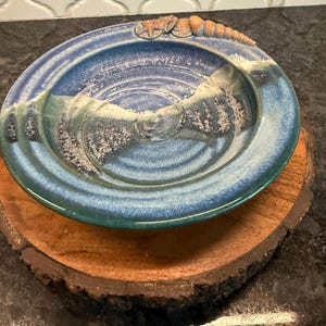 May include: A handcrafted ceramic plate with a blue and green glaze, featuring a raised design and seashell accents. The plate rests on a round, wooden base with a natural edge, creating a rustic, coastal aesthetic.