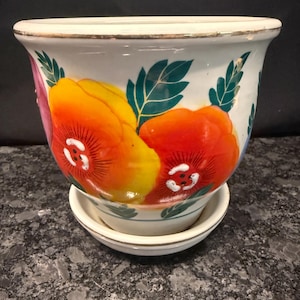 May include: A white ceramic planter with a matching saucer, featuring a hand-painted floral design with large yellow and orange poppy-like flowers, dark green leaves, and a thin gold-toned metallic rim around the top edge of the pot and saucer.