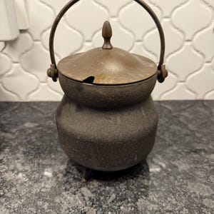 May include: A small, antique-style metal pot with a lid and handle. The pot is dark gray with a bronze-colored lid and handle. It has a small, decorative knob on top of the lid and three small feet.