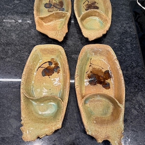 May include: Four handmade ceramic serving dishes. Each dish is oval-shaped with a small, attached bowl and a decorative floral design. The dishes are primarily tan with brown and green accents. The edges have a textured, organic appearance.