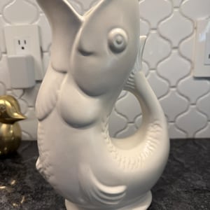 May include: A white ceramic pitcher in the shape of a fish. The pitcher has a large open mouth, a tail that forms a handle, and detailed scales. The pitcher is a decorative item for the kitchen or dining room.