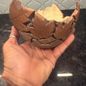 May include: A handcrafted ceramic bowl with a leaf design. The bowl is brown with a textured surface and leaf imprints. The interior is a lighter tan color. The bowl is held in a hand.