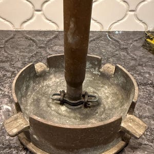 May include: A vintage ashtray with a central cylindrical holder. The ashtray is made of metal with a weathered, aged appearance. It features a circular base with four resting points for cigarettes.