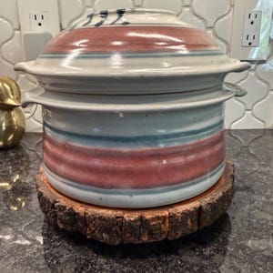 May include: A ceramic cooking pot with a lid, featuring horizontal bands of red and blue on a white background. The pot sits on a rustic wooden coaster. The lid has the number "192" on it.