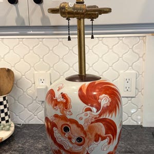May include: A decorative ceramic table lamp with a white base and red and orange painted design. The lamp has a brass stem and a brown wooden base. The lamp is approximately 20 inches tall.