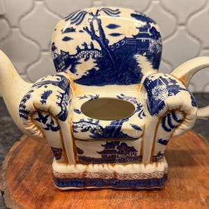 May include: A cream-colored teapot shaped like an armchair, featuring a blue and white Willow pattern design. The teapot has a spout, handle, and a central opening. It sits on a wooden surface.