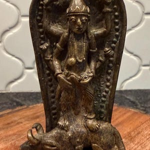 May include: A bronze-colored statue depicting a deity riding an animal, possibly a bull. The figure has multiple arms and is adorned with jewelry. The statue is mounted on a base and set against a decorative backdrop. The overall style suggests a religious or cultural artifact.