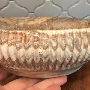 May include: A decorative bowl with a unique, textured design. The bowl features a wavy, ridged pattern in shades of beige, brown, and cream. The rim is smooth, and the bowl is set on a wooden base. This item is suitable for home decor.