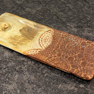 May include: A rectangular ceramic tray with a textured surface. The tray features a color palette of brown, tan, and cream. The right side has a bubbly texture, while the left side has a smooth finish with a small, raised circular feature.