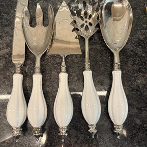 May include: A set of five serving utensils with white handles and silver-toned metal heads. The set includes a knife, fork, pie server, slotted spoon, and serving spoon. The handles have a bulbous shape with a ribbed design.