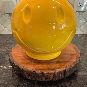 May include: A large, glossy yellow smiley face sculpture with oval eye cutouts and a curved smile. It sits atop a round, natural wood slice with a rough bark edge. The sculpture is a cheerful decorative item.