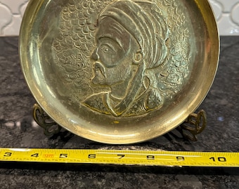 May include: A brass tray with an embossed design of a man's face with a turban. The tray is on a stand with two decorative feet.