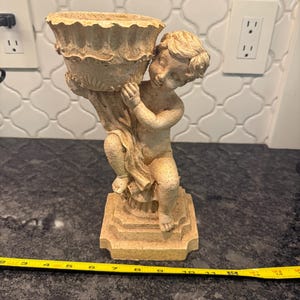 May include: A beige cherub statue holding a decorative urn. The statue is on a square base and measures approximately 10 inches in length. The cherub has curly hair and is smiling. The urn has a ruffled edge.
