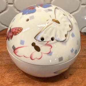 May include: A white, spherical ceramic container with butterfly and geometric designs. The container features pink, blue, and gold accents. The top has several small holes. The container is sitting on a wooden surface.