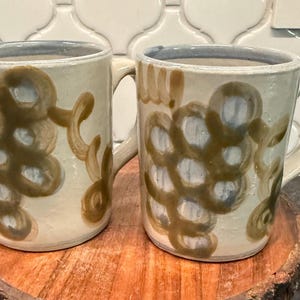 May include: Two ceramic mugs with handles. The mugs are off-white with a blue rim and feature a brown, abstract design. They are displayed on a round, wooden coaster. The mugs are suitable for coffee, tea, or other beverages.