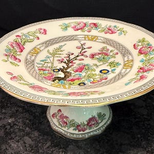 May include: A ceramic pedestal cake stand featuring a cream-colored surface with a central floral motif of pink blossoms and green leaves. The rim and base are decorated with a black Greek key pattern and gold trim, with additional floral accents.