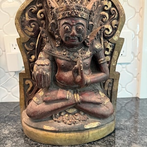 May include: A carved wooden statue of a seated figure with a detailed headdress and ornate gold accents. The statue is primarily reddish-brown with gold highlights, showcasing intricate carvings and a serene expression. The figure is in a meditative pose.