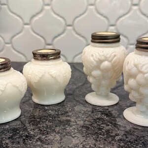 May include: Four white glass salt and pepper shakers with silver-colored lids. Two shakers have a butterfly design, and two have a grape cluster design. The shakers are arranged on a dark countertop.