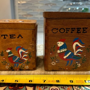 May include: Two wooden canisters with painted roosters and the words "TEA" and "COFFEE" on the front. The canisters are brown with red, blue, and white accents.