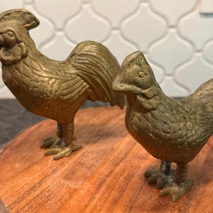 May include: Two brass rooster and hen figurines. The rooster stands tall with a detailed comb and tail feathers, while the hen is smaller and more compact. Both are positioned on a round wooden base, showcasing a rustic, farmhouse aesthetic.