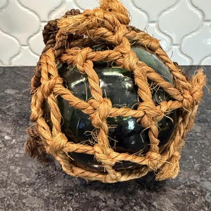 May include: A dark green glass float encased in a woven rope net. The rope is a natural tan color, and the float has a glossy finish. The float is a decorative item, possibly for nautical-themed decor.