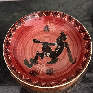 May include: A red ceramic bowl with a black silhouette of a person dancing. The bowl has a white rim with a black and white geometric pattern. The bowl is sitting on a wooden base.