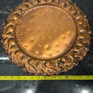 May include: A copper-colored decorative plate with an ornate, raised floral design around the rim. The plate measures approximately 15 inches in diameter. The center of the plate has a series of small, evenly spaced indentations.