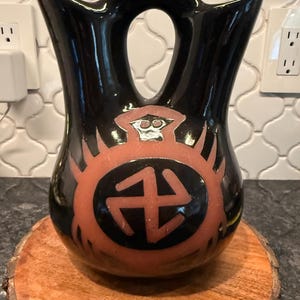 May include: Black ceramic vase with a unique shape, featuring a handle and a central opening. The vase has a reddish-orange design with geometric shapes. It sits on a round wooden base, showcasing a blend of textures and colors.