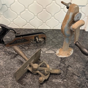 May include: Three antique woodworking tools are displayed. A black and brown hand plane with a black handle, a rusty metal crank-operated drill, and a metal tool with a handle and a blade are visible. The tools are on a dark surface.